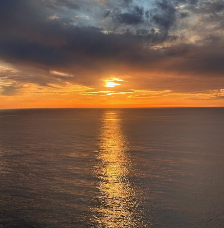 A vivid ocean sunset viewed from a high coastal cliff, with golden light reflecting in a straight path across calm water beneath dramatic storm clouds.