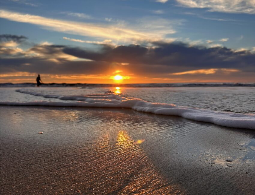A wave rolls onto wet sand at sunset, reflecting golden light, with a lone silhouette standing at the water's edge