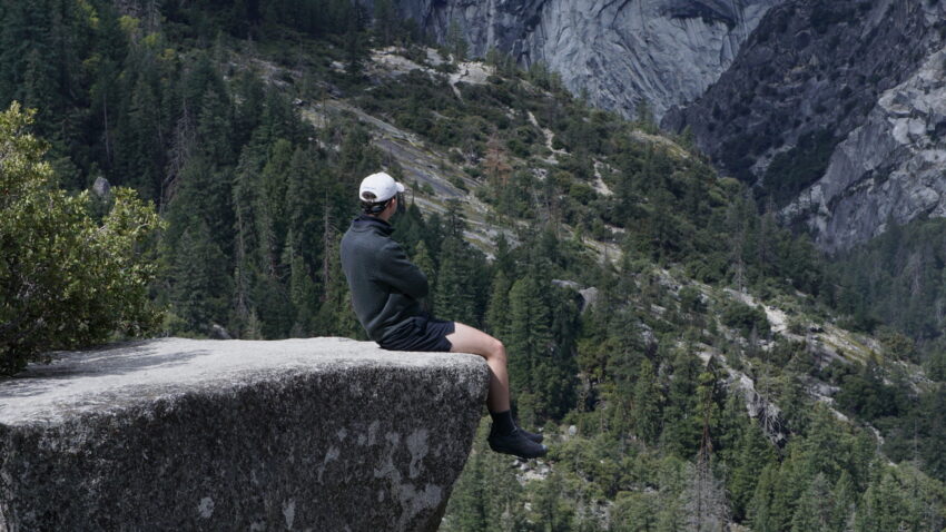 A figure sits on the edge of a granite ledge above Nevada Falls, looking out over the Yosemite valley below.