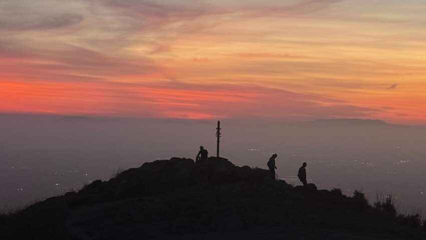 Three silhouetted figures stand at the summit of Mission Peak at dusk, beside the summit pole, with an orange sunset band and the hazy lights of the South Bay below.