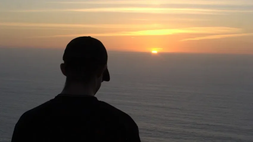 A silhouetted figure looks out over the ocean at sunset from a coastal cliff.